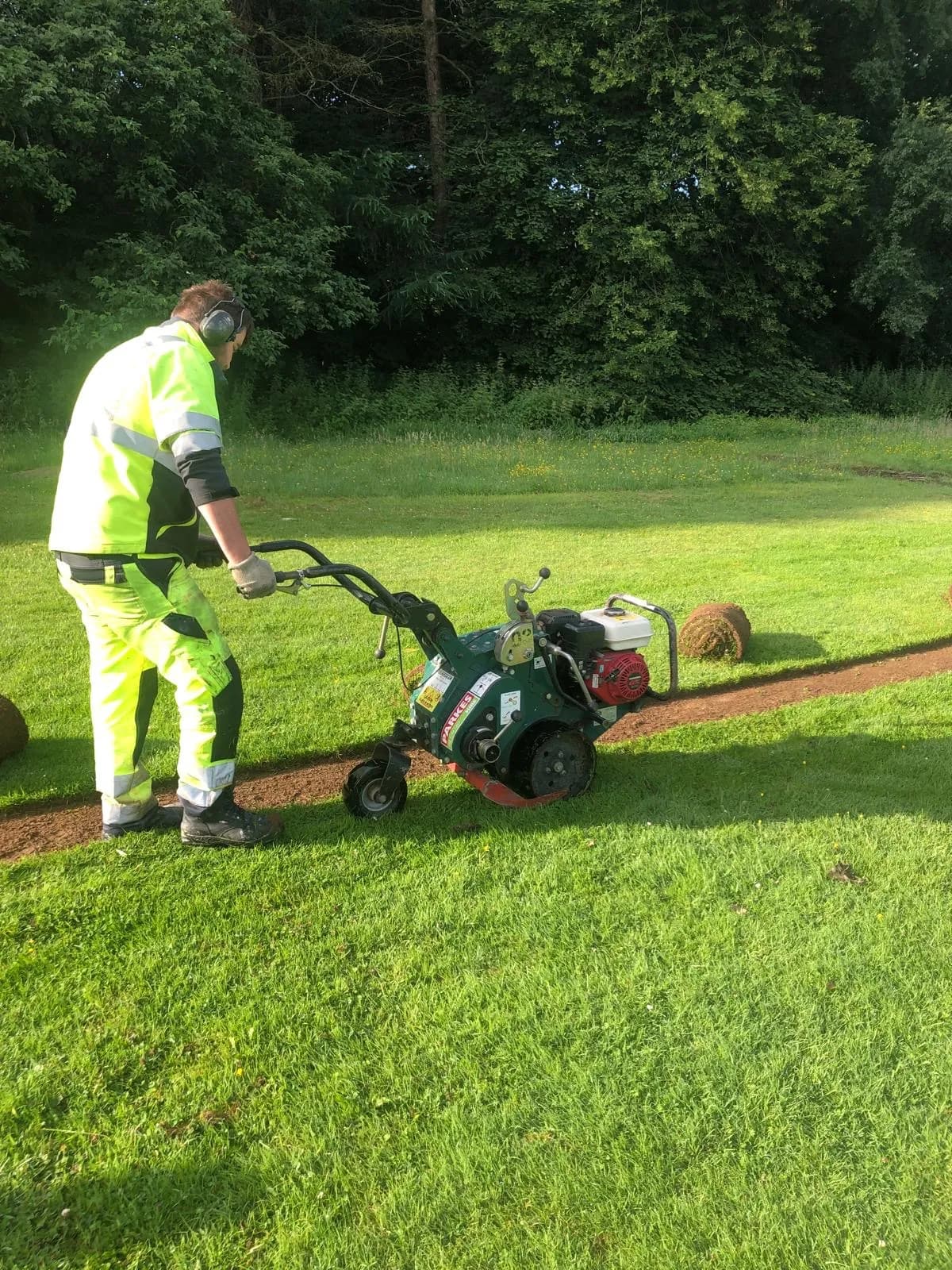 A person in neon-yellow work clothes with ear protection operates a sod cutter on a grassy lawn with trees in the background, creating a strip of removed grass and revealing the soil underneath.
