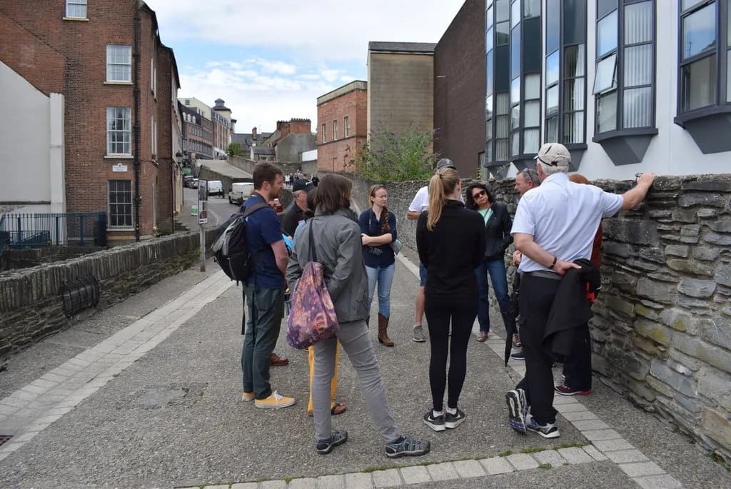 A group of people are gathered on a stone pathway with brick buildings and a street in the background.